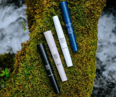 Vier Dolomia Mascaras in Schwarz, Beige, Weiß und Dunkelblau auf bemoostem Stein, darunter Waterproof Long Lasting Intense Black Lashes.